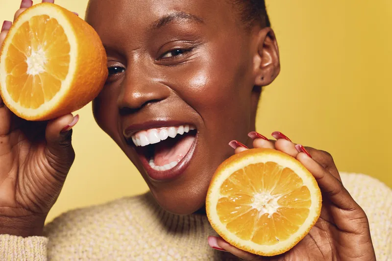 Smiling black woman with dark skin holding two halves of a fresh orange fruit close to her face. The yellow background and natural light highlight healthy skin and joyful mood.