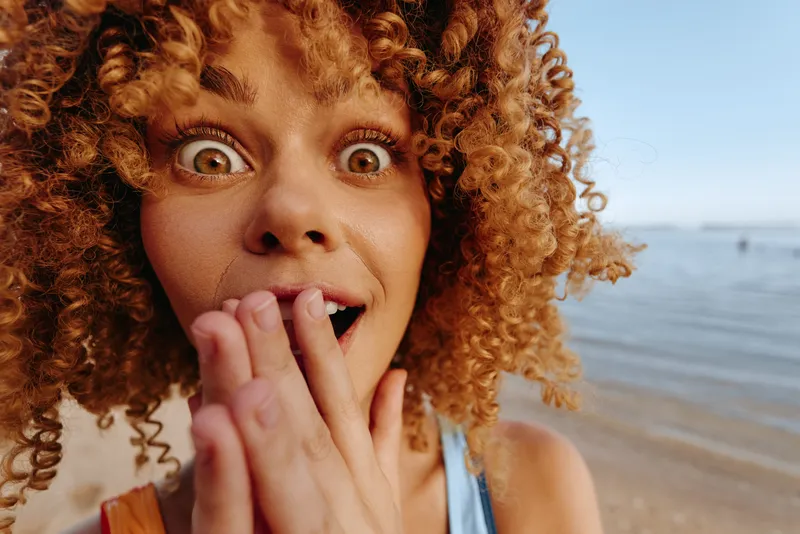 Joyful close up of a surprised woman with curly hair at the beach, bright sunlight, candid expression, playful mood, warm tones, ocean background, summer vibe in the foreground.