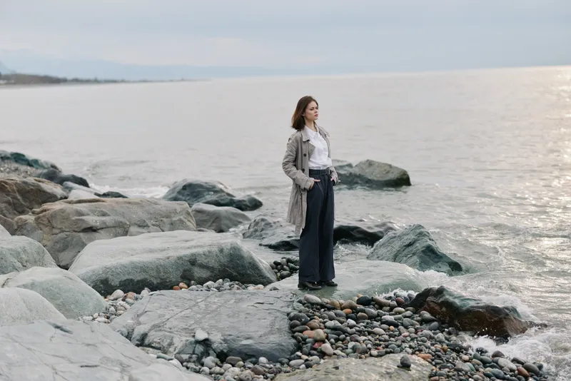 A thoughtful woman stands on rocky shore facing calm sea under cloudy sky. She wears a long coat and dark pants, reflecting on peaceful nature and solitude by the water.