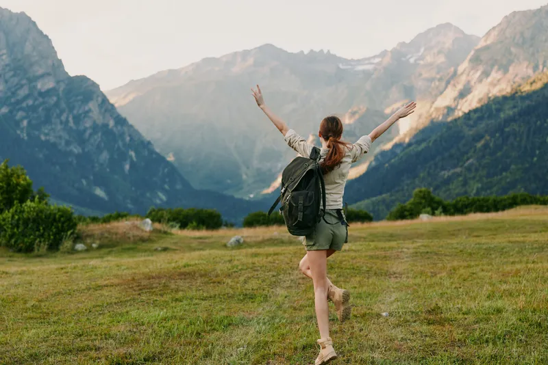 A young woman with a backpack raises her arms while walking on a grassy field surrounded by mountains under a clear sky. Concept for lifestyle, travel, and freedom.