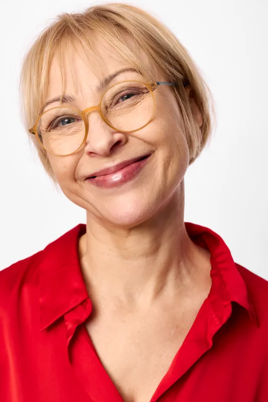 Senior woman, blond hair, wearing glasses, smiling, studio portrait, red shirt, friendly expression, clean white background, calm mood, closeup, indoor