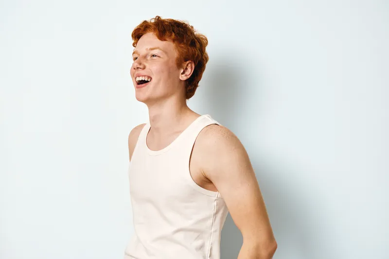 Young redhead man laughing with joy wearing white sleeveless shirt isolated on white background. Casual happy male expression with ginger hair and freckled face in studio.