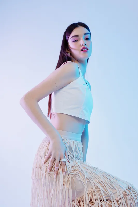 Young woman in a white crop top and fringed skirt poses confidently in a studio with soft blue lighting. Concept of beauty, fashion, and modern style.