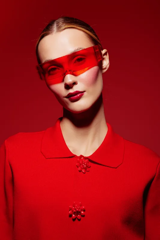 Portrait of a stylish woman wearing red sunglasses and a red sweater with decorative buttons. The red background and lighting create a bold, modern fashion concept.