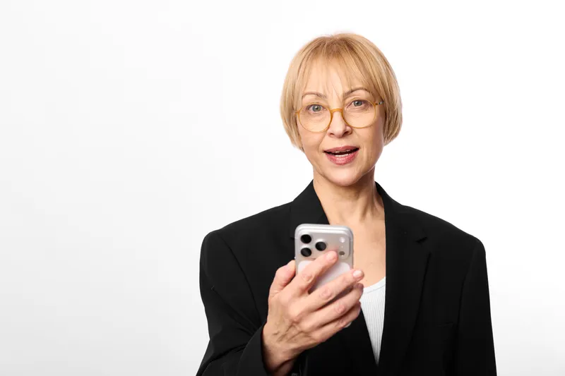 Senior woman with glasses holds a smartphone in a studio portrait, looking at camera with calm focus. Business communication concept about technology use, everyday.