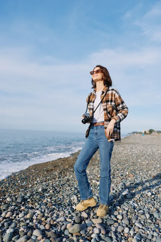 Woman on a rocky beach with a camera, wearing casual plaid shirt and jeans, enjoying coastal scenery under a sunny sky with sea breeze