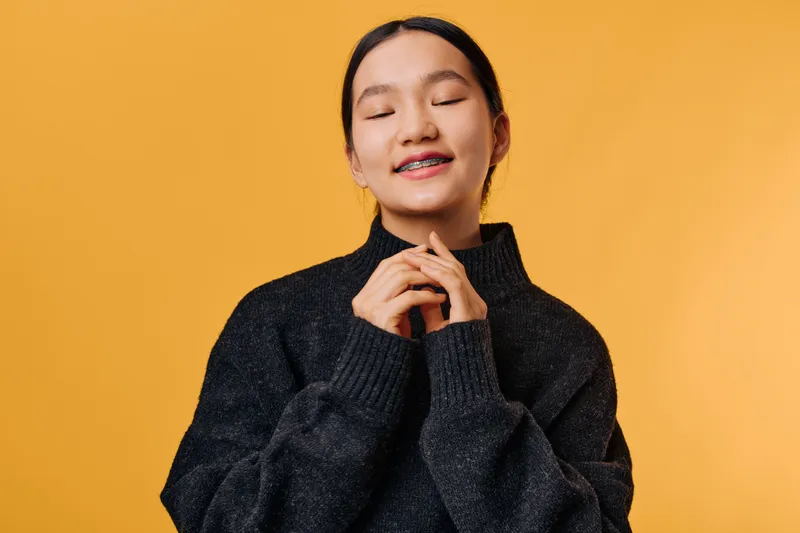 Young woman with braces smiling with closed eyes wearing dark sweater against orange background. Relaxed person with hands close to face showing calm expression and peaceful mood.
