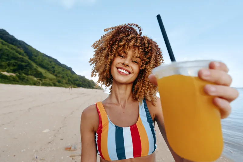 A smiling young woman with curly hair holds a cold orange drink with a black straw on a sunny beach with hills and clear sky in the background.