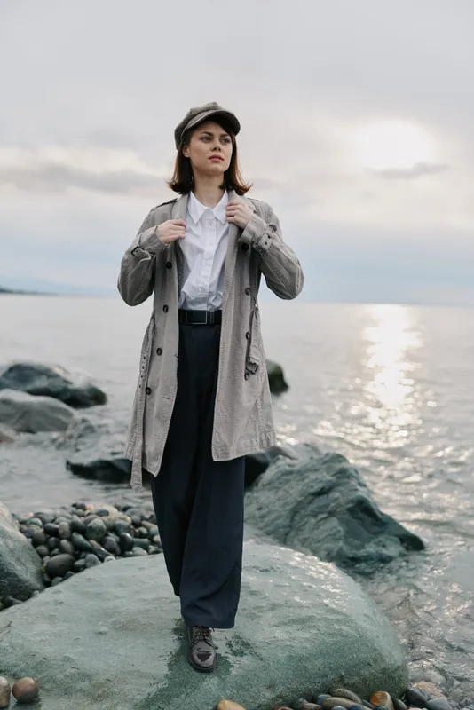 Young woman in a plaid coat and cap stands on a rocky shore near the sea during cloudy sunset. Fashion concept with casual autumn outfit and natural landscape background.