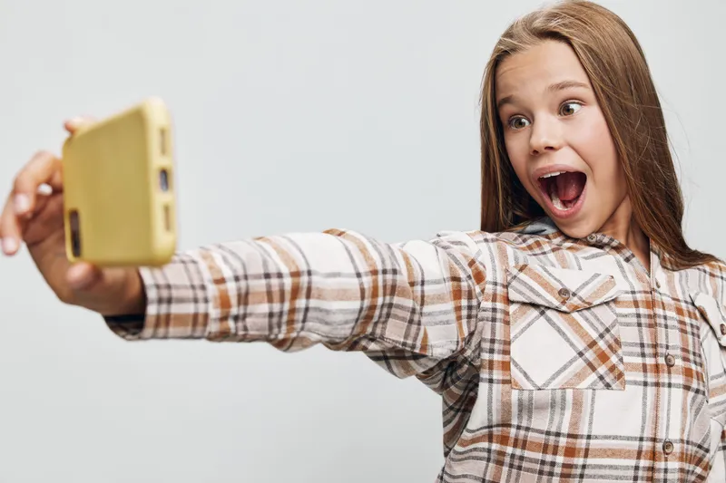Girl taking selfie with phone smiling happy and surprised wearing plaid shirt on light background in studio with long hair and casual style.