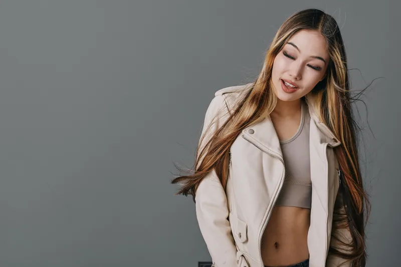 Young woman with long hair and closed eyes wearing beige jacket and crop top posing against gray background for fashion, beauty and casual style photo shoot session.