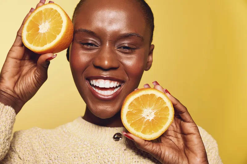 Joyful African woman with dark skin holds two halves of orange fruit near face smiling against yellow background. Concept of healthy lifestyle, vitamin, happiness, and natural beauty.
