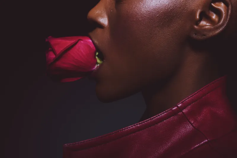 Close-up of a dark skinned African woman with smooth skin holding a red rose in her mouth. She wears a red leather jacket against a dark background. Concept of beauty and sensuality.
