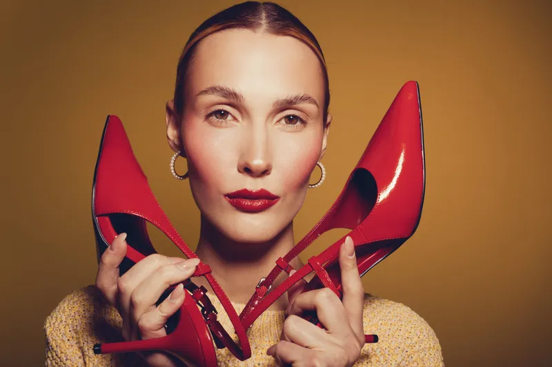 Close-up of confident woman holding red high heels near face on brown background. Concept of fashion, style, elegance, and beauty with bold makeup and accessories.