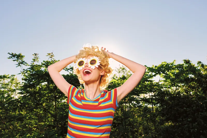 Smiling woman with curly hair wearing flower glasses and a striped shirt in bright sunlight. Outdoor setting conveys playful summer vibes, carefree style, and bold color.
