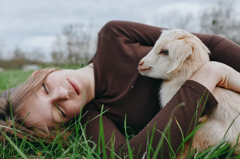 Relaxed woman lying on grass with a goat kid, sharing a gentle moment outdoors. Natural light, warm tones, lifestyle scene highlighting companionship, relaxation, nature, and calm wellbeing in a