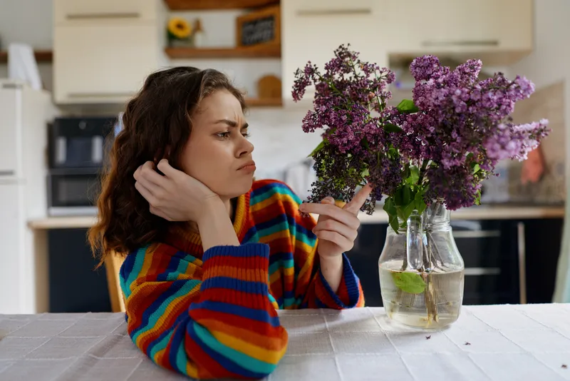 A woman in a striped sweater sits at a kitchen table, gazing at a vase of purple flowers with a thoughtful expression, capturing a home lifestyle moment of calm and beauty.