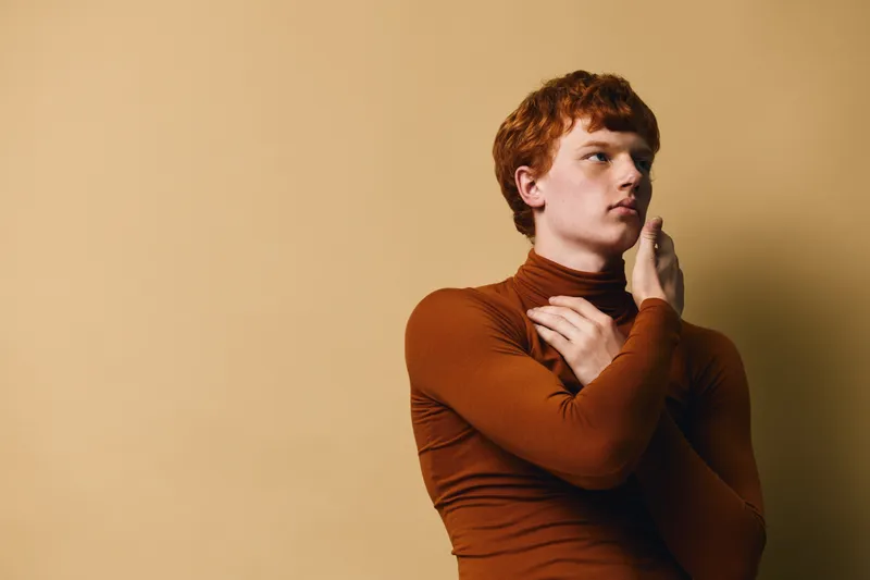 Young man with red hair posing in brown turtleneck sweater against beige background for fashion, style, portrait and expression in studio lighting.