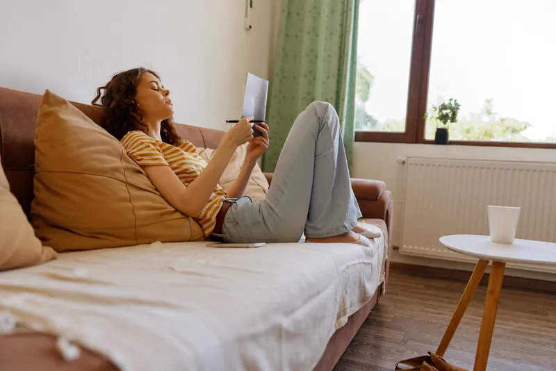 Young woman relaxing on a sofa at home, browsing a tablet with a gentle smile, cozy living room scene, soft natural light