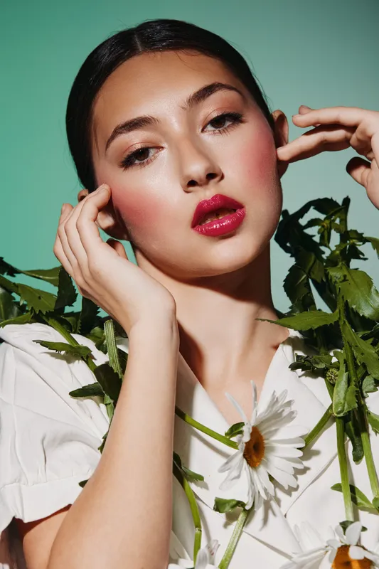 Close-up of a fashion woman with glossy red lips and natural makeup posing with fresh daisies on green background. Concept of beauty creative emotion and modern style.