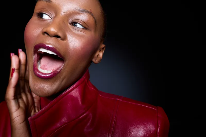 Close-up of a happy black woman with dark skin wearing a red leather jacket, smiling with mouth open and hand near face. Studio shot on black background. Concept of joy and fashion.