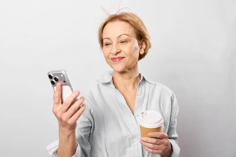 Woman smartphone coffee portrait smiling older indoors casual beverage communication, mature woman holds mobile phone and coffee cup, smiling toward camera, relaxed morning routine at home on white