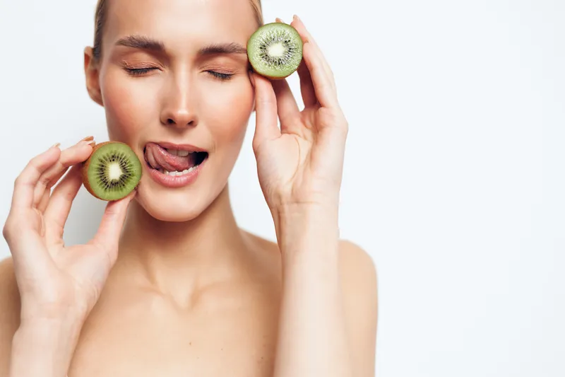 A playful woman holds fresh kiwis near her face, lips parted in a light smile as she bites into the fruit. Bright studio lighting highlights healthy skin and a skincare vibe.