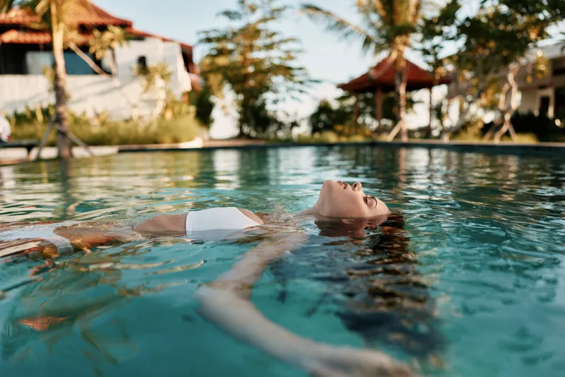 Relaxed woman floating on back in outdoor pool, serene expression, sunny day, clear water, tropical resort background, peaceful lifestyle moment, calm and refreshing scene
