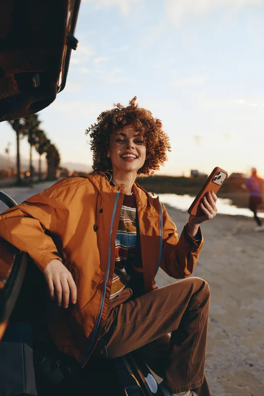 Smiling woman outdoors in warm sunshine, curly hair and casual jacket, holding smartphone while sitting by parked van, travel lifestyle mood, soft breeze, golden street background