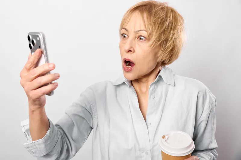 Older woman, surprised, holding smartphone and skincare cream jar, studio portrait on white background, skin care moment, copy space, soft lighting, neutral mood