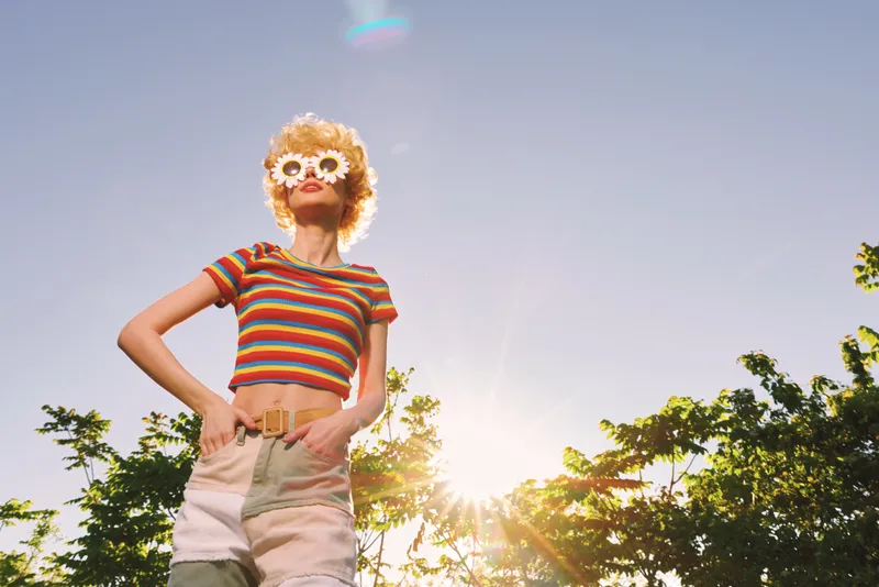 Woman in striped crop top and high waisted shorts poses outdoors in bright sun, retro film color mood, casual lifestyle vibe, playful expression, sunny sky and green trees in the background