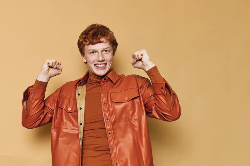 Young man with red hair smiling and raising fists in celebration wearing brown shirt and orange jacket standing against plain beige background in studio.
