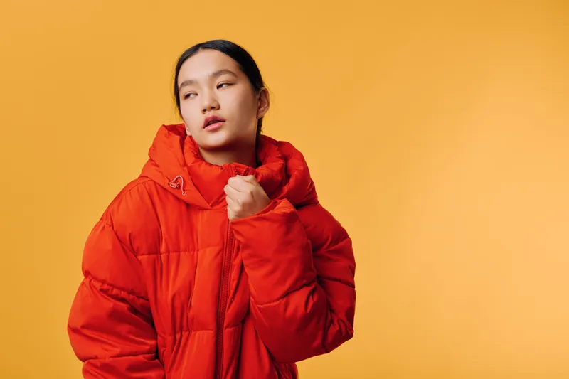 Young woman in red warm jacket standing against yellow background looking to side with thoughtful expression and casual pose for winter fashion or lifestyle.