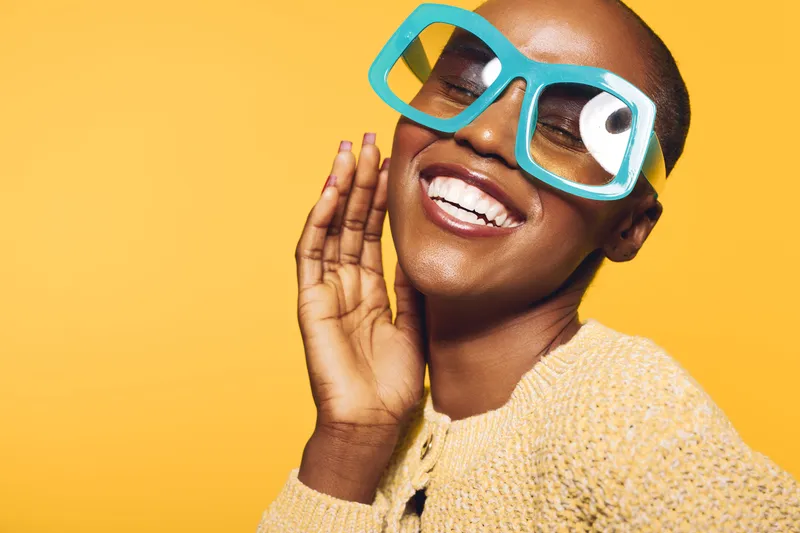 Smiling dark skinned African woman with short hair wearing large blue glasses and yellow sweater poses against yellow background. Concept of joy, fashion, and positive lifestyle.