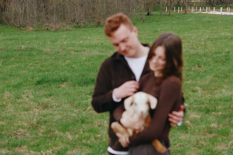 Couple outdoors, affection moment with a puppy in a sunlit field, casual wear, warm tones, soft breeze, natural backdrop, lifestyle scene capturing joy and connection and togetherness
