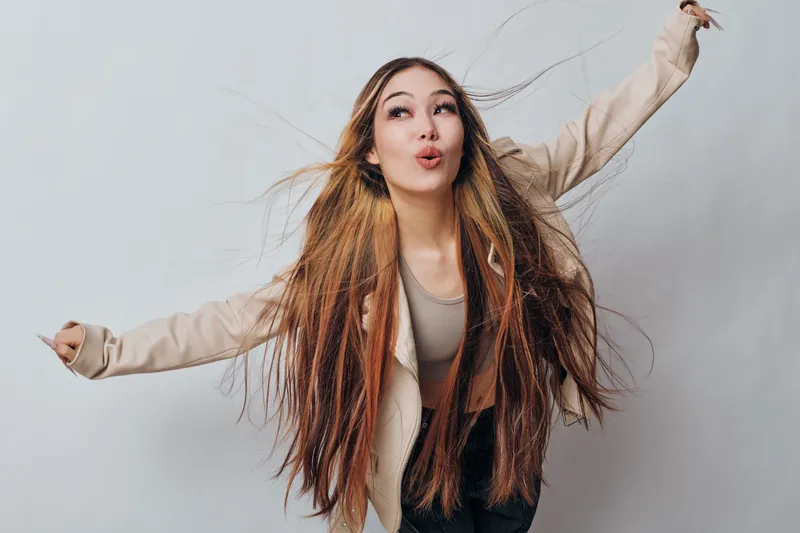 Woman with long hair posing with excitement and energy against gray background. Casual style with beige jacket and expressive face showing fun and motion.