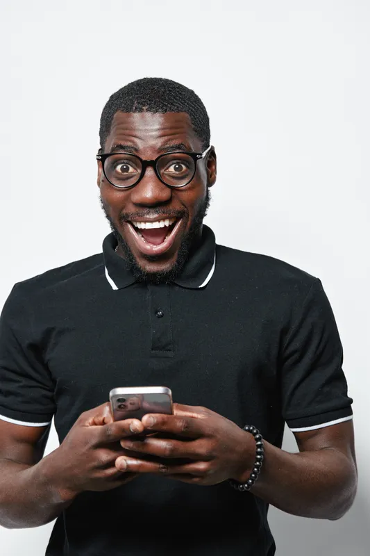 African American man with dark skin and glasses smiling excitedly while holding smartphone in black polo shirt on white background. Happy, casual, technology use.