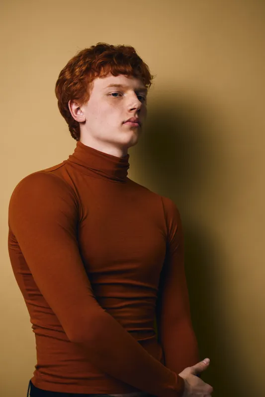 Young man with red hair and pale skin wearing brown turtleneck standing against plain beige background with thoughtful confident expression and crossed arms.
