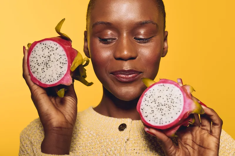 Close-up of a happy African woman with dark skin holding two halves of a dragon fruit against a yellow background. Concept of healthy lifestyle, exotic fruit, and natural beauty.