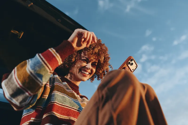 Smiling woman with curly hair holding a phone for a selfie outdoors, playful portrait moment in summer sunlight, casual clothing, lifestyle focus, relaxed friendly mood
