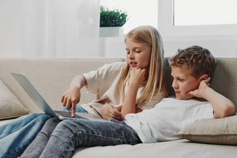 Two children lying on a couch using a laptop together in a bright room. Focused boy and girl explore digital content, learning or playing in a cozy home environment with natural light.