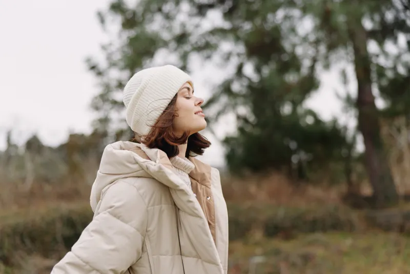 Woman in a puffy winter coat and knit beanie stands outdoors in a park, enjoying the fresh air and soft light, a serene moment of warm comfort and quiet reflection.