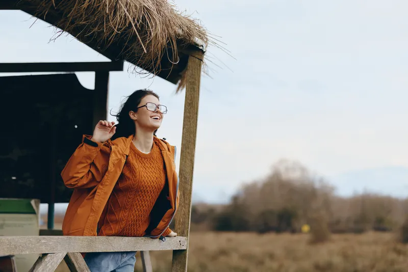 Smiling woman with glasses, outdoor lifestyle, orange jacket and denim jeans, leaning near wooden shelter with straw roof, autumn landscape background, soft daylight, cheerful smile