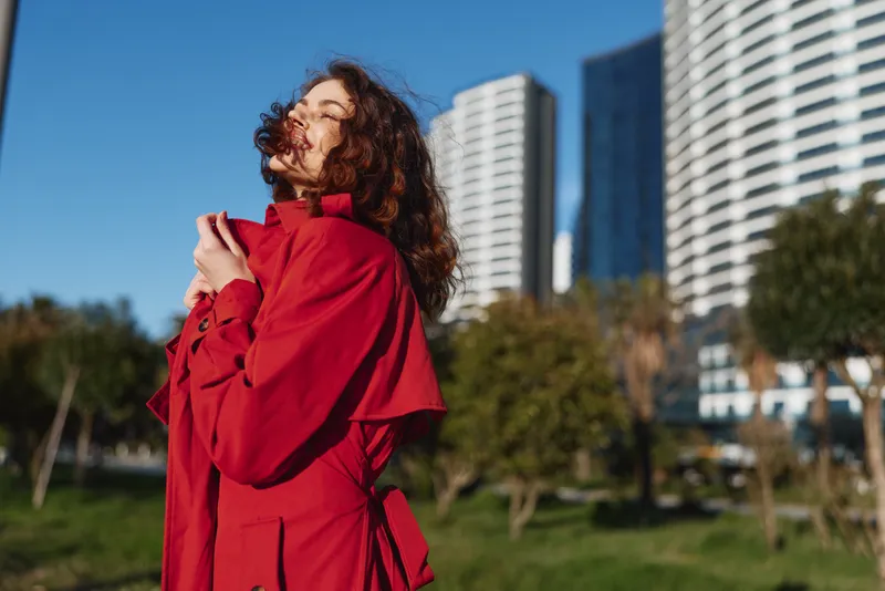 Young woman in red coat enjoys fresh air outdoors with closed eyes, standing in sunny park near modern buildings, feeling calm and relaxed in urban environment.