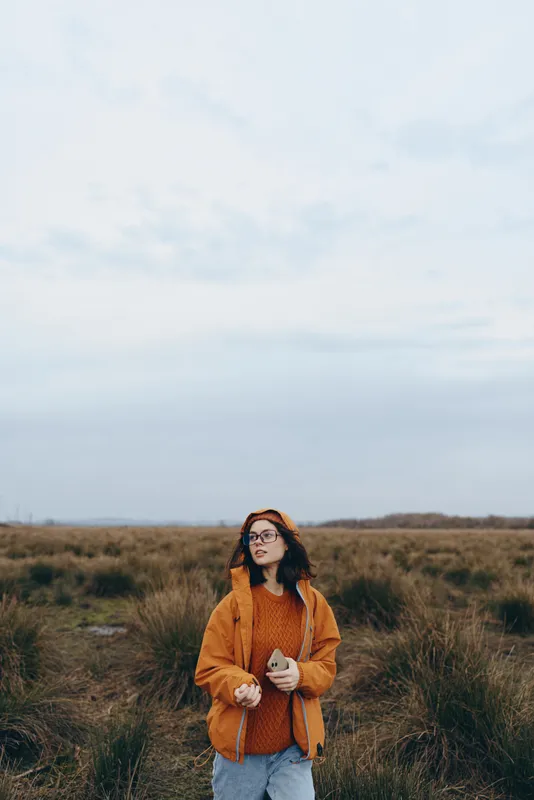 Woman lifestyle outdoors, autumn field with tall grass, woman in orange jacket and casual sweater, gentle breeze, relaxed stance, looking at camera, soft daylight, natural warm colors, calm mood