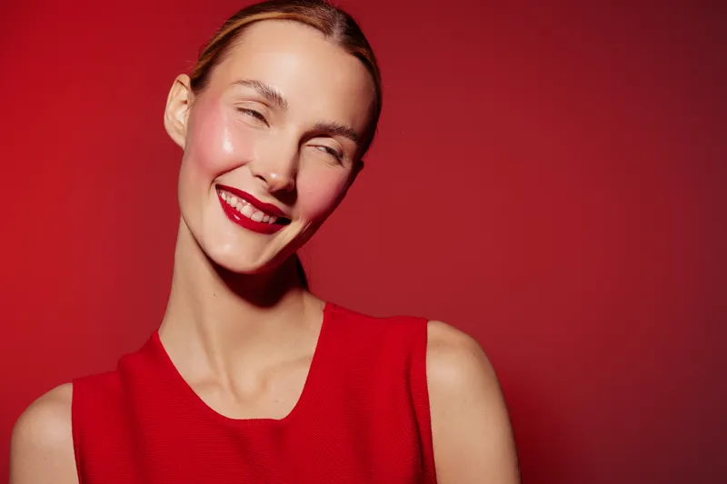 Smiling young woman with red lipstick and red sleeveless top poses against red background. Concept of beauty, fashion, makeup, and confident female portrait in studio lighting.