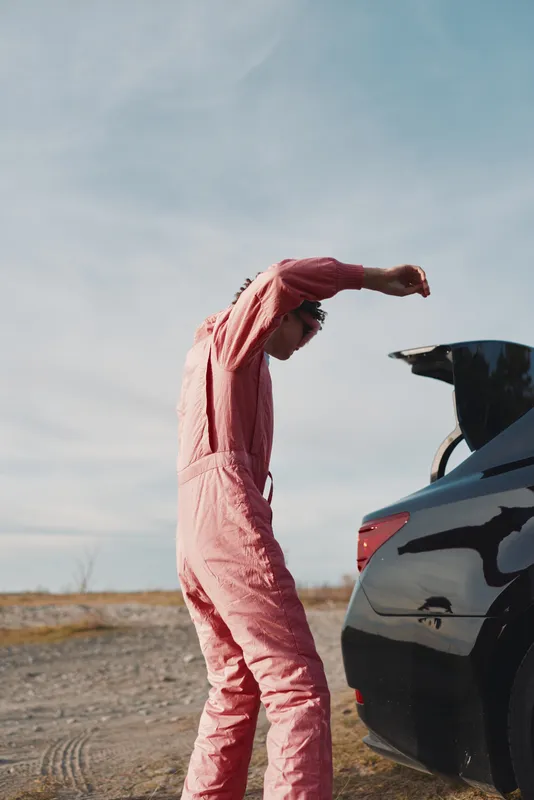 Outdoor scene with a person in pink workwear stretching beside an open car trunk in a desert landscape, preparing for loading or unloading gear in bright daylight.