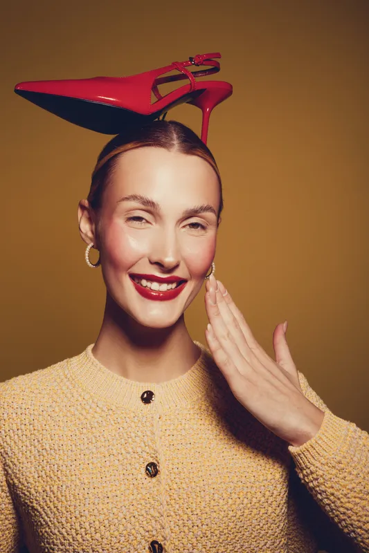 Smiling woman with red high heel shoe on head wears yellow textured jacket and gold earrings. Studio portrait on brown background with playful and fashionable concept.