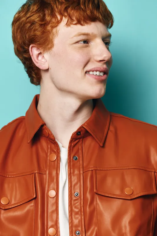Redhead young man portrait in brown leather jacket smiling and looking side on blue background. Casual style with natural expression and modern fashion for male model photography.