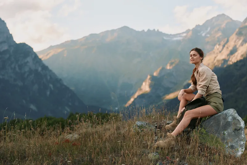 Woman sits on a rock in a mountain meadow, enjoying the view. Outdoor lifestyle scene highlighting nature, solitude, and calm.
